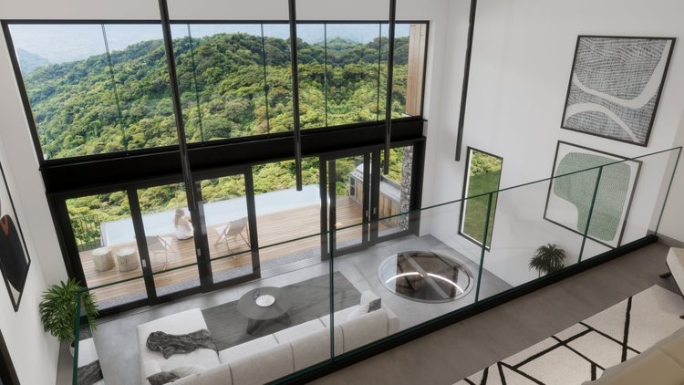 Living room with glass railing overlooking a large window with a view of lush green hills. The room has a white sofa with a throw blanket, a round coffee table, and wall art. Outside on the balcony, a woman sits on a chair near a barbecue grill, enjo