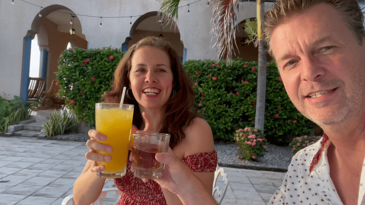 Smiling woman holding a yellow drink and a small glass with a darker beverage, with a man partially visible taking a selfie together outdoors in a sunny, tropical setting with lush green plants and a building with arches in the background.