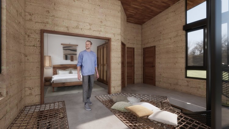 A man is standing in a rustic wooden room with large windows, inside a house or cabin. In the adjacent bedroom, there is a bed with white bedding, and a woven wall hanging. The room features wood panel walls, wooden sliding doors, and a patterned rug