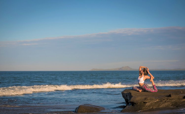 A woman practicing yoga on a rocky beach at sunset, with the ocean and distant islands in the background.