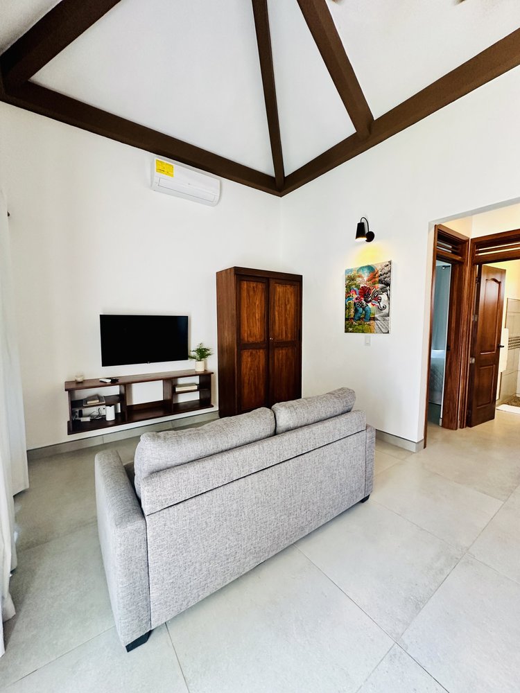 Living room with a beige sofa, wall-mounted TV, wooden cabinet, colorful painting, and doorway leading to bathroom.