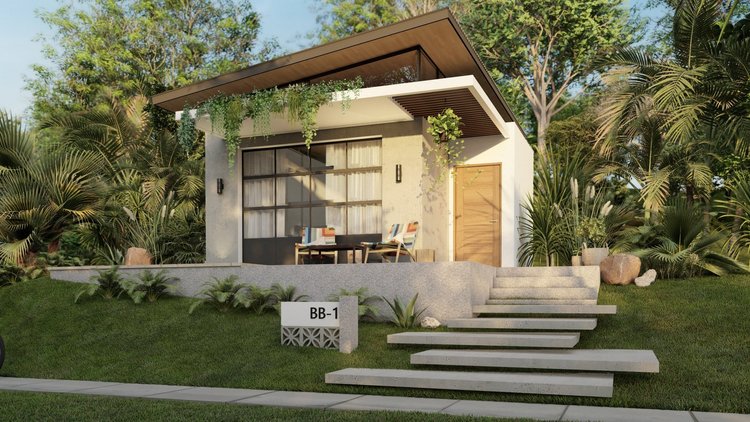 Modern small house with white walls, wooden door, large glass windows, and a slanted roof surrounded by lush green trees and plants. There's a staircase leading up to the entrance and an outdoor seating area with striped chairs.