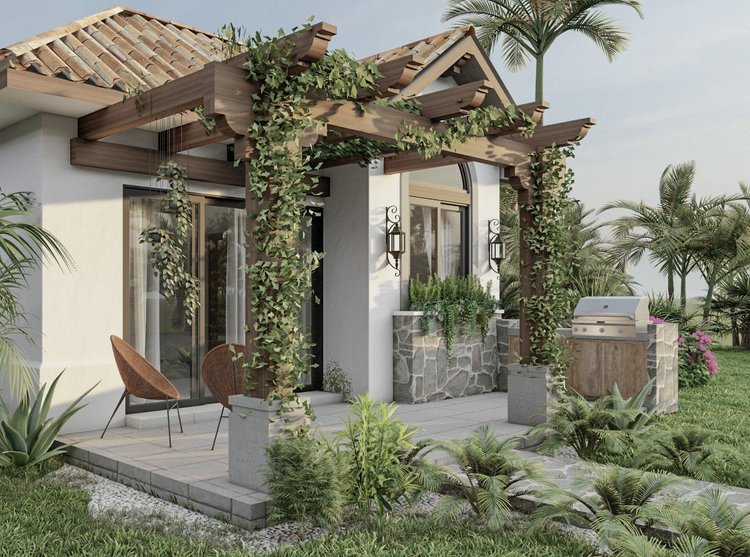 Front porch of a house with a wooden pergola draped with ivy, two wicker chairs on a tiled patio, lush tropical plants, a stone water feature, and a built-in barbecue, surrounded by greenery and palm trees.
