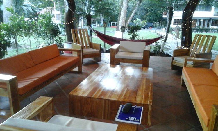Indoor seating area with wooden furniture, including orange and beige couches and chairs, a large wooden coffee table, and a small side table. Outside, a hammock and trees are visible through large windows, with string lights hanging from the trees.