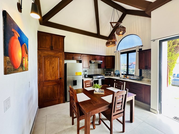 A bright kitchen with wooden cabinets, a dining table with four chairs, a stainless steel refrigerator, a stove, and a window with a view outside. Woven hanging pendant lights and artwork on the wall add decor.